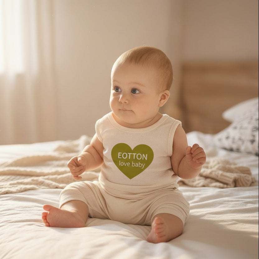 A baby wearing a sleeveless beige tank top and matching harem pants, sitting on a bed.