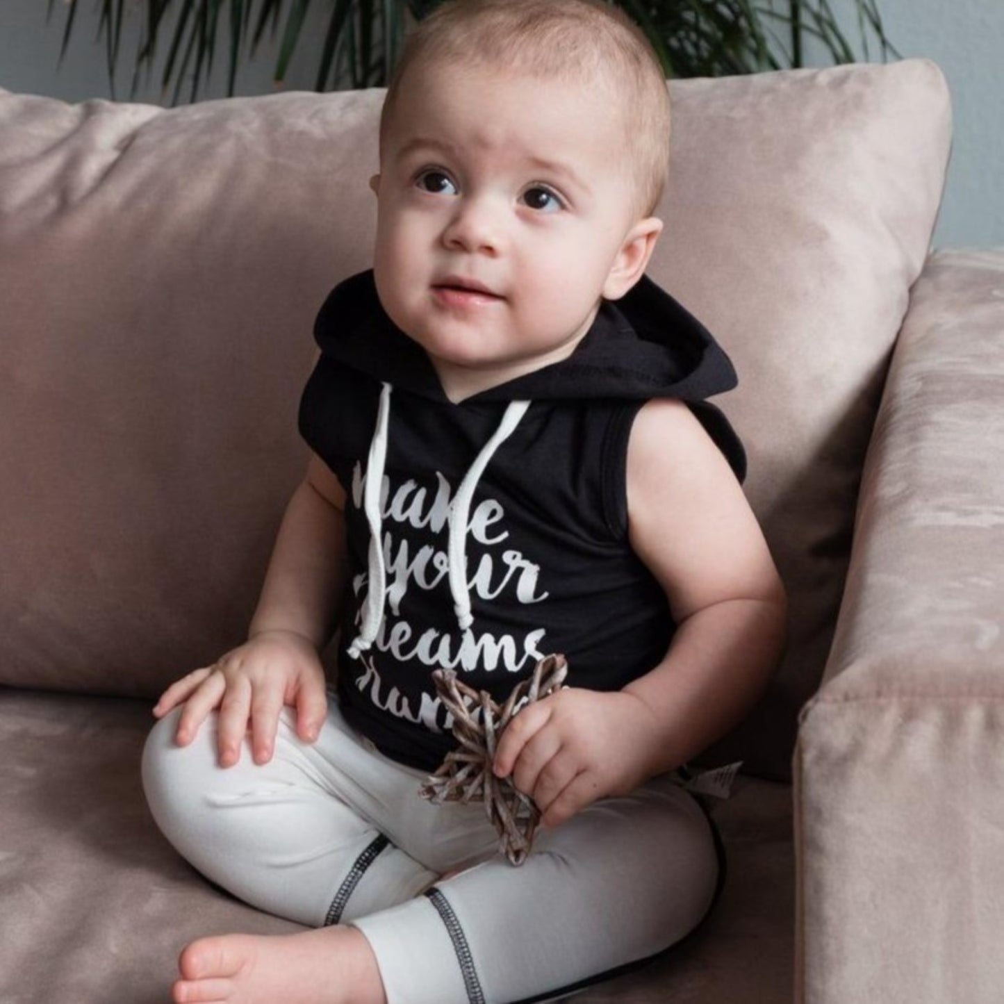 Cute active baby sitting on a couch wearing the black motivational sleeveless hooded vest and white organic cotton pants.