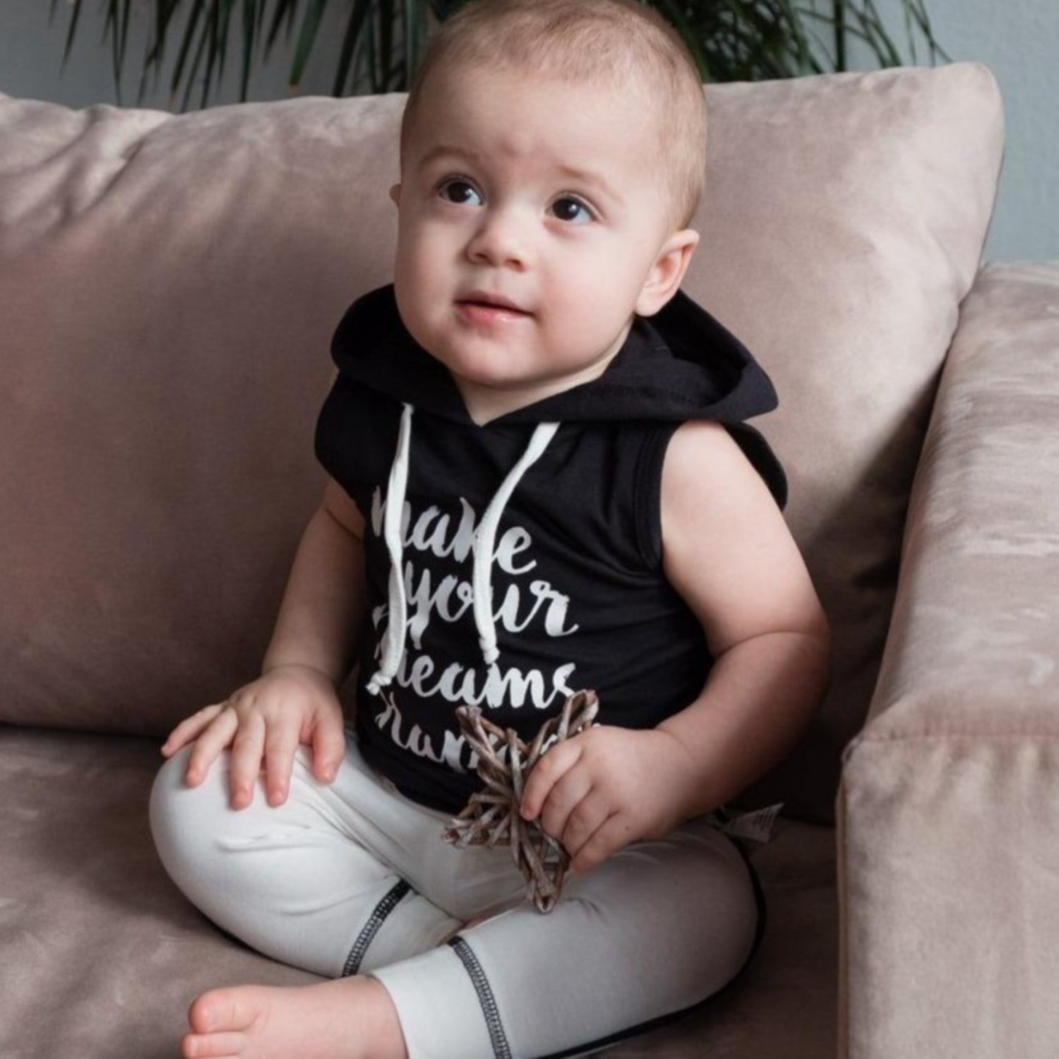 Cute active baby sitting on a couch wearing the black motivational sleeveless hooded vest and white organic cotton pants.