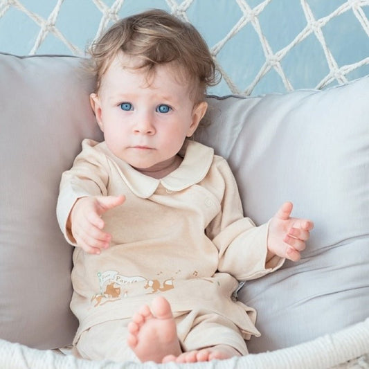 A baby girl wearing a two piece play suit made from natural cream certified organic combed cotton featuring a side snap wrap shirt with a Peter Pan collar and matching pants