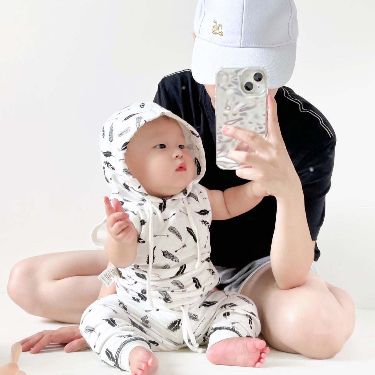 Little baby sitting down wearing the monochrome feather pattern organic sleeveless hoodie and active toddler jogger suit.